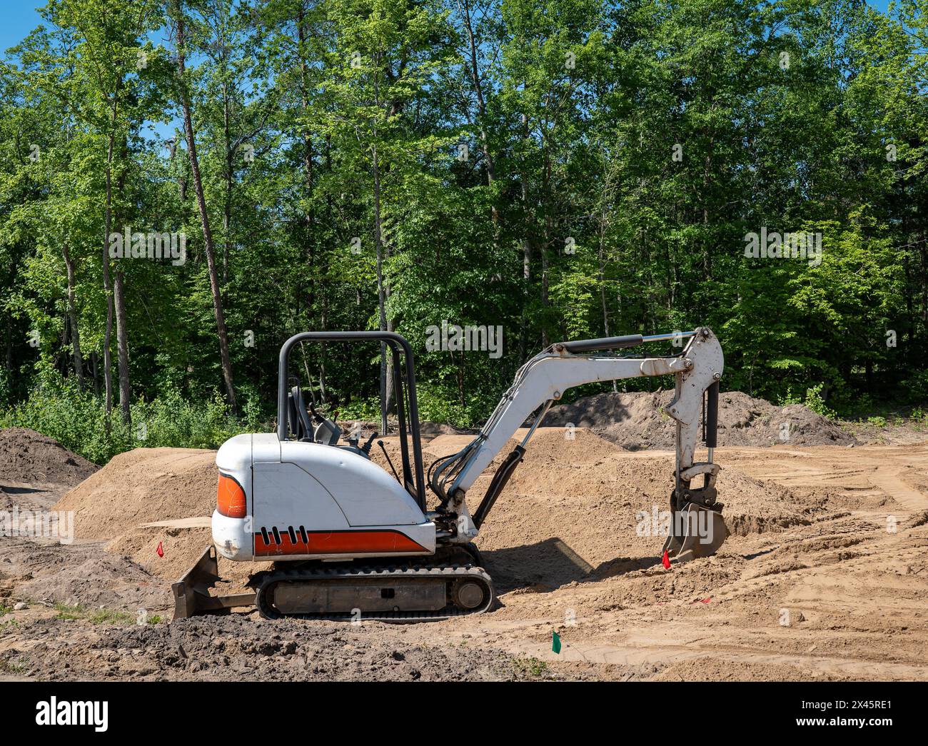 Un miniescavatore con paletta idraulica si trova sullo sporco in un nuovo lotto di costruzione della casa, con alberi sul retro del sito. Foto Stock