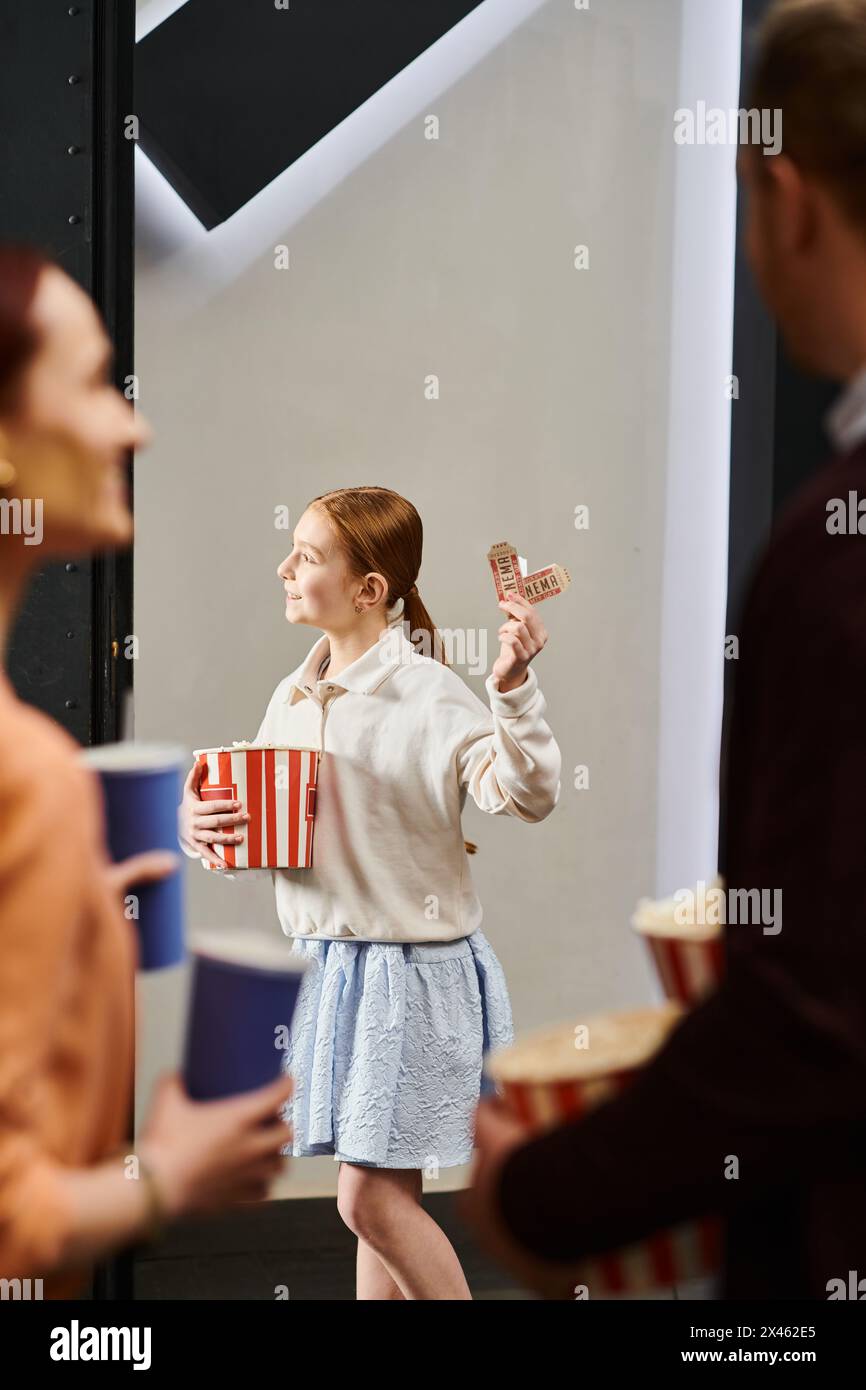 Una ragazza tiene con gioia una scatola di popcorn e i biglietti, godendosi una serata al cinema con la famiglia. Foto Stock