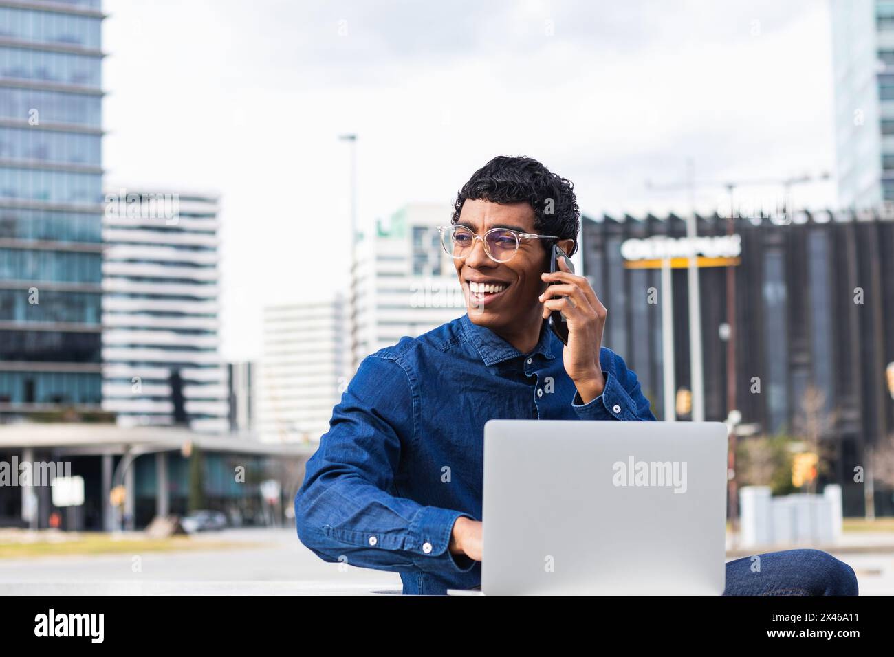 Adulto etnico allegro dipendente remoto maschile che ha una conversazione su smartphone mentre si lavora su netbook in città nelle giornate di sole Foto Stock