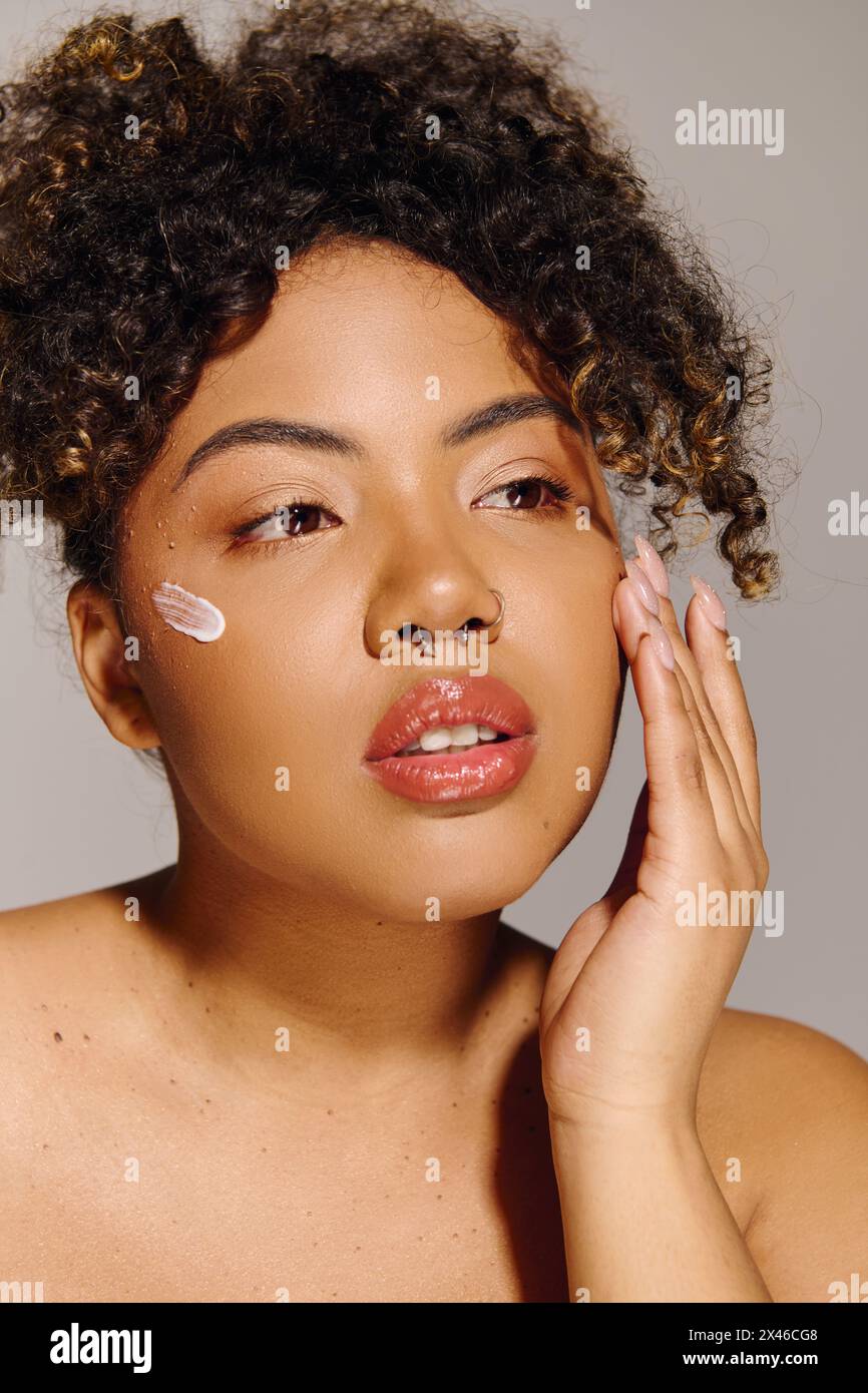 Una bella giovane donna afroamericana con capelli ricci che mostra una sorprendente crema bianca sulla guancia in un ambiente da studio. Foto Stock