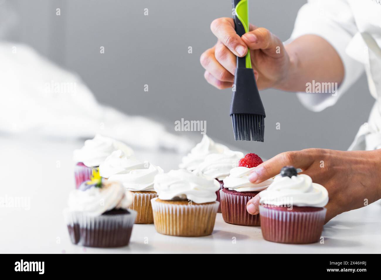Panettiere irriconoscibile con spazzola in silicone che sparge sciroppo dolce sulle bacche mentre prepara i cupcake vegani in cucina Foto Stock