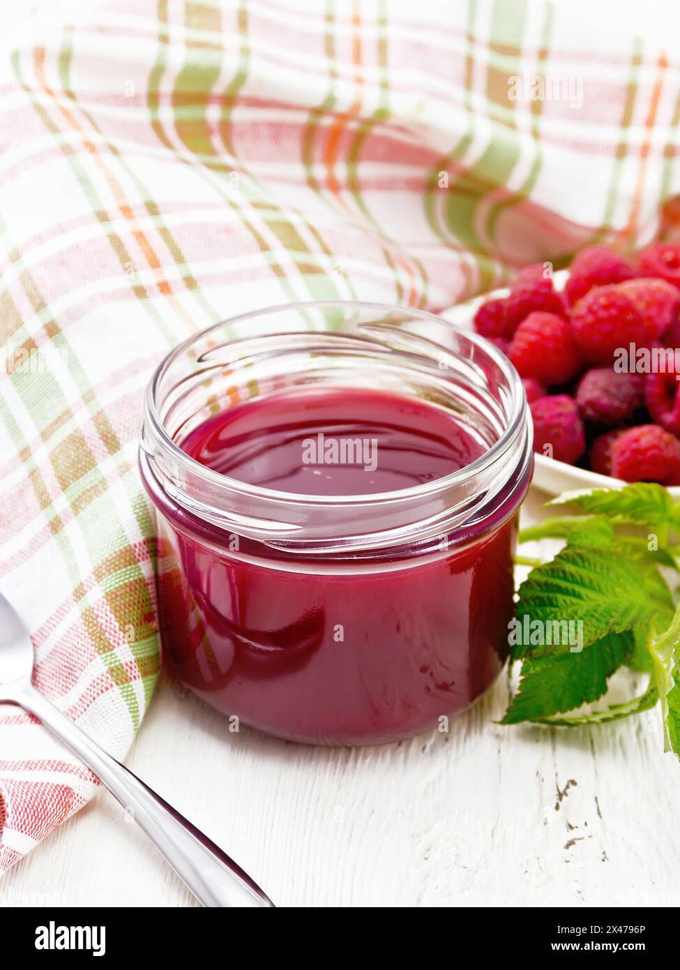 Marmellata di lamponi in vaso di vetro, frutti di bosco nel recipiente, foglie verdi, asciugamano e cucchiaio su sfondo in legno Foto Stock