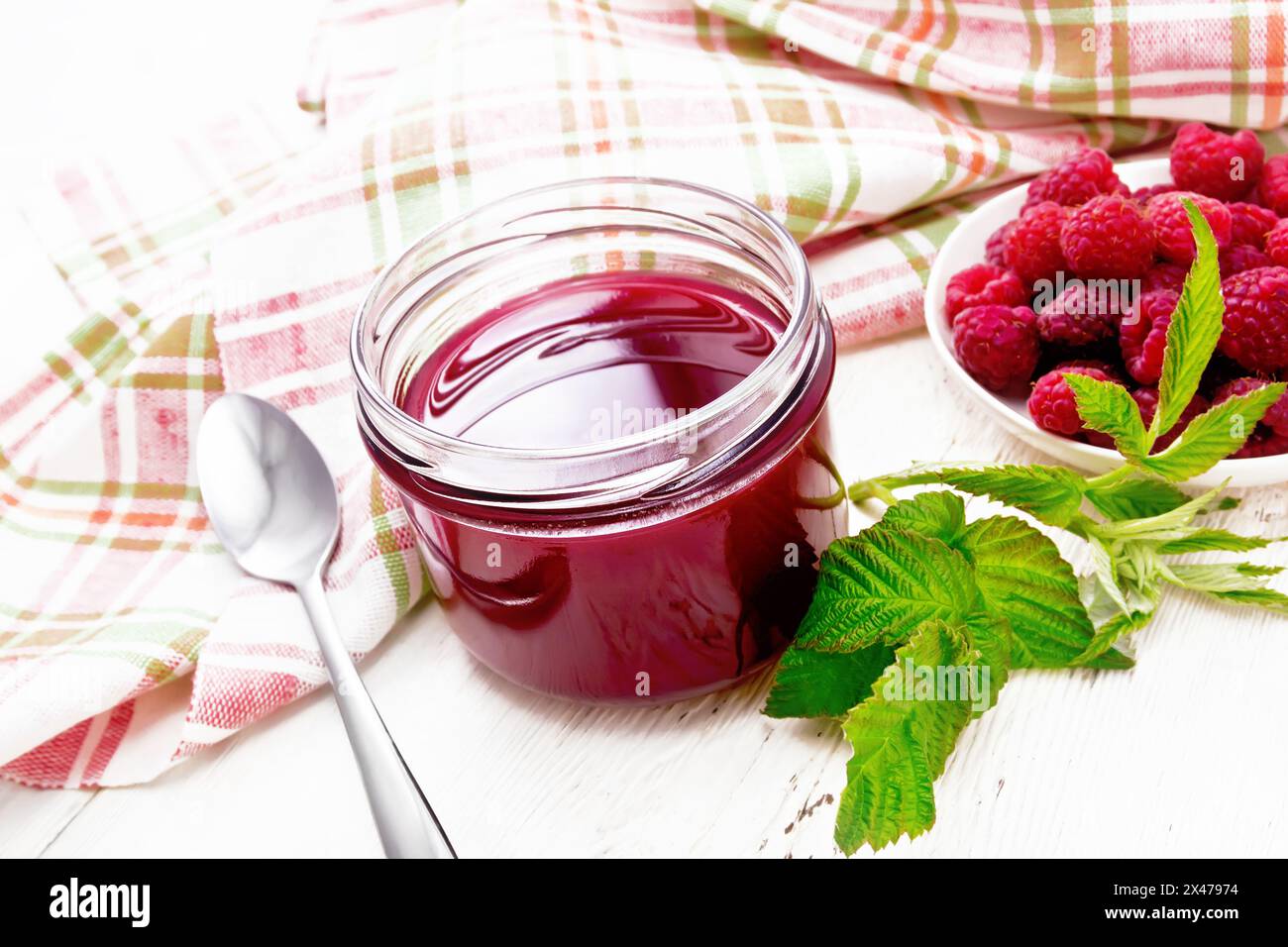 Marmellata di lamponi in vaso di vetro, frutti di bosco nel recipiente, foglie verdi, asciugamano e cucchiaio sullo sfondo di un bordo di legno bianco Foto Stock