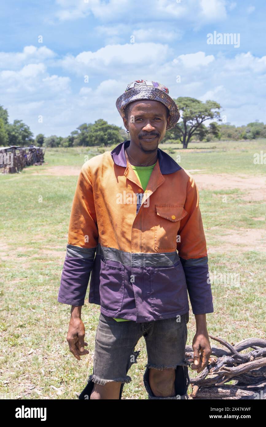 giovane operaio africano del villaggio, indossa abiti da lavoro arancioni, vende legna da ardere in un campo verde Foto Stock