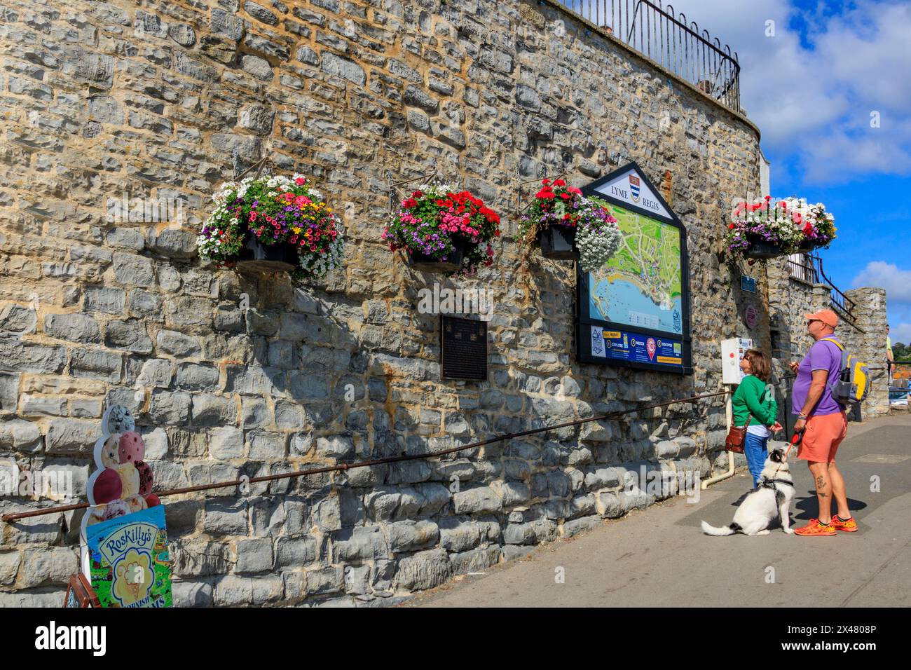 Visitatori che studiano una grande mappa della città circondata da attraenti e colorati cesti appesi a Lyme Regis, Dorset, Inghilterra, Regno Unito Foto Stock