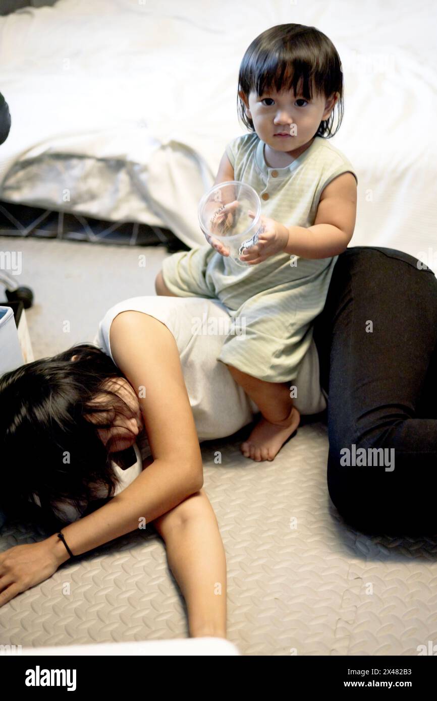 La madre esausta giace sul pavimento mentre il figlio gioca su di lei. Le madri esauste soffrono di depressione postpartum. Foto Stock