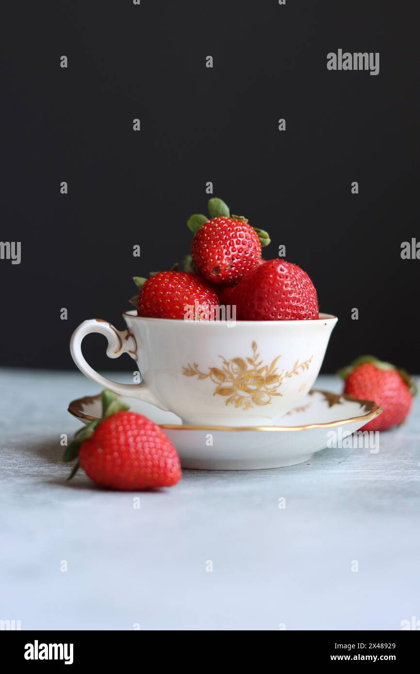 Fragole in una piccola tazza di porcellana bianca e dorata su sfondo scuro con spazio per il testo. Foto STILL Life con stoviglie retrò e frutta estiva. E Foto Stock