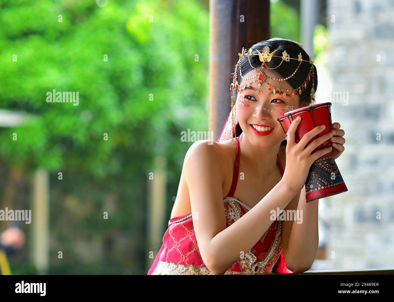 Bella ragazza cinese con un sorriso radioso in tradizionale costume rosso delle minoranze etniche e copricapo con un tamburo, Nanjing, provincia di Jiangsu, Cina Foto Stock