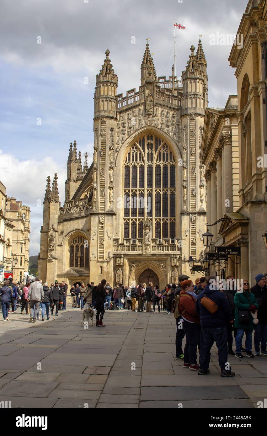 25 aprile 24 la storica Abbazia di Bath nel Somerset, Inghilterra, con turisti che camminano nell'Abbey Churchyard in una bella mattinata primaverile. La bandiera di St Georges e' in alto Foto Stock