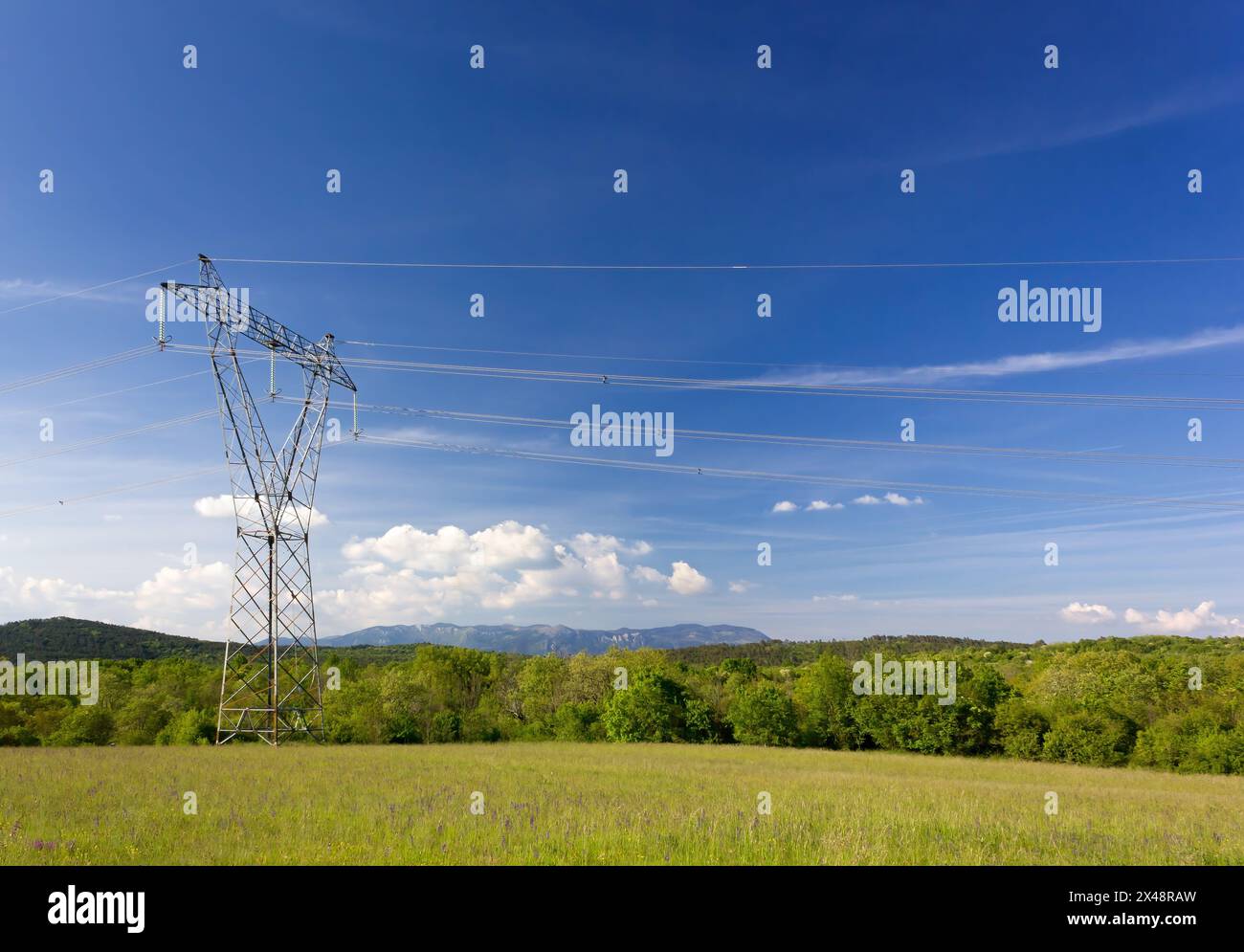 Campo verde su un cielo blu con un pilone di elettrcittà in primo piano e colline e montagne sullo sfondo Foto Stock