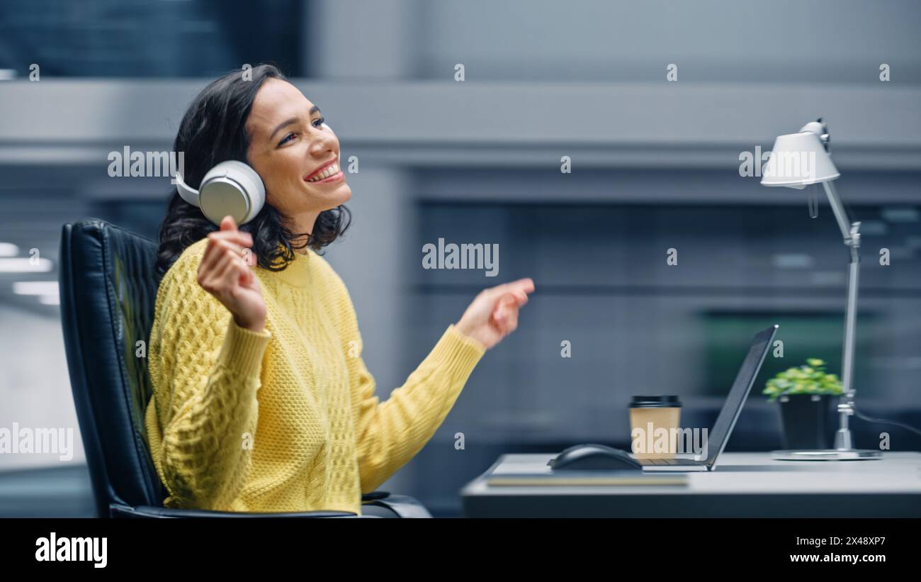 Ufficio moderno: Donna d'affari ispanica sorridente seduta alla scrivania che lavora al computer, indossa le cuffie, ascolta musica, balli. E' felice, si diverte, divertirsi. Sfondo sfocatura movimento Foto Stock