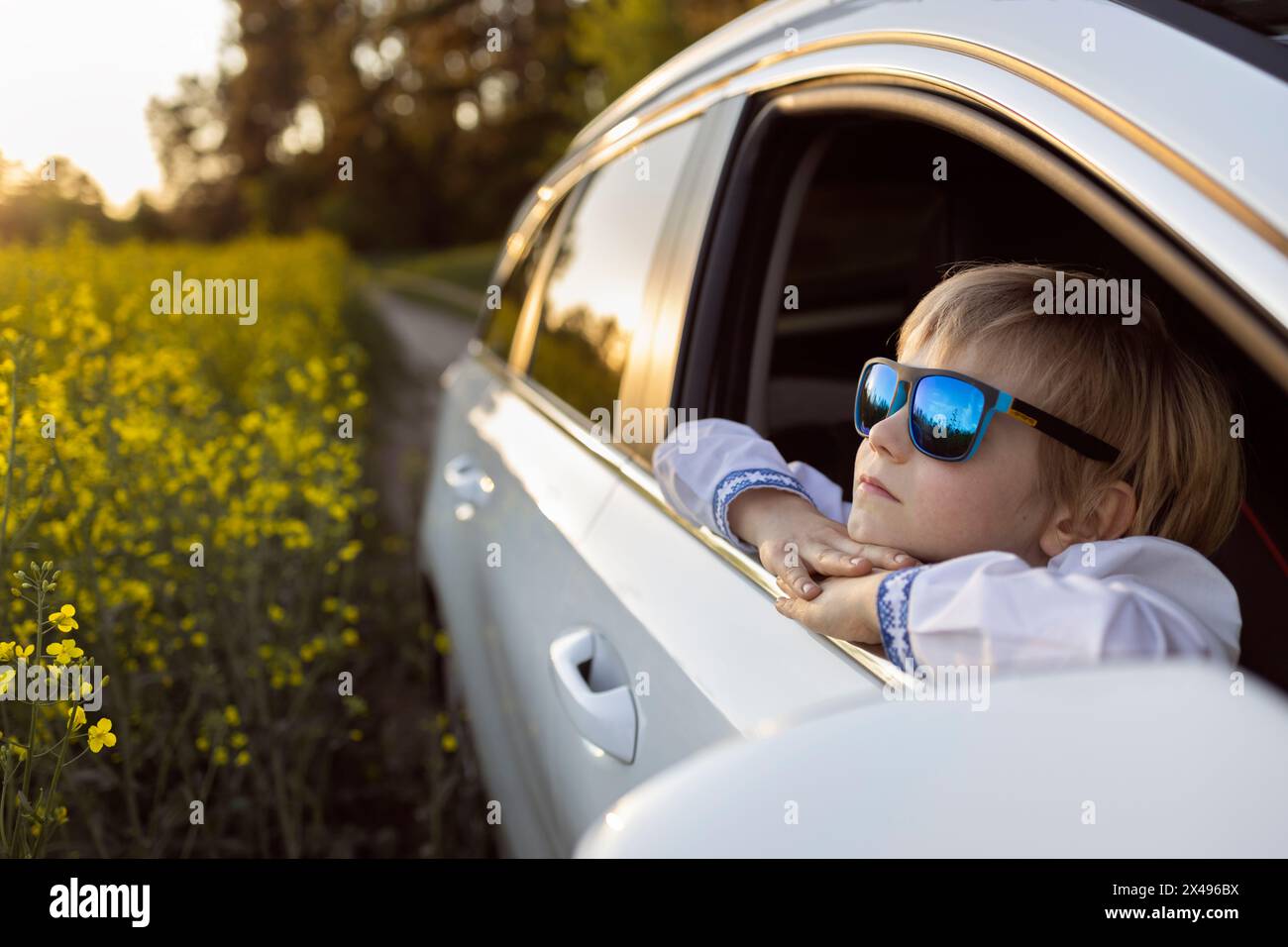 la faccia felice di un bambino con gli occhiali da sole blu guarda fuori dal finestrino dell'auto aperto e si guarda intorno con interesse. viaggi in auto con la famiglia in estate Foto Stock