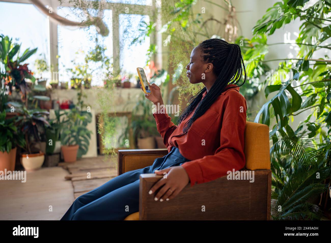 Una donna afroamericana rilassata e contenta siede su una poltrona con smartphone circondato da piante interne. Foto Stock