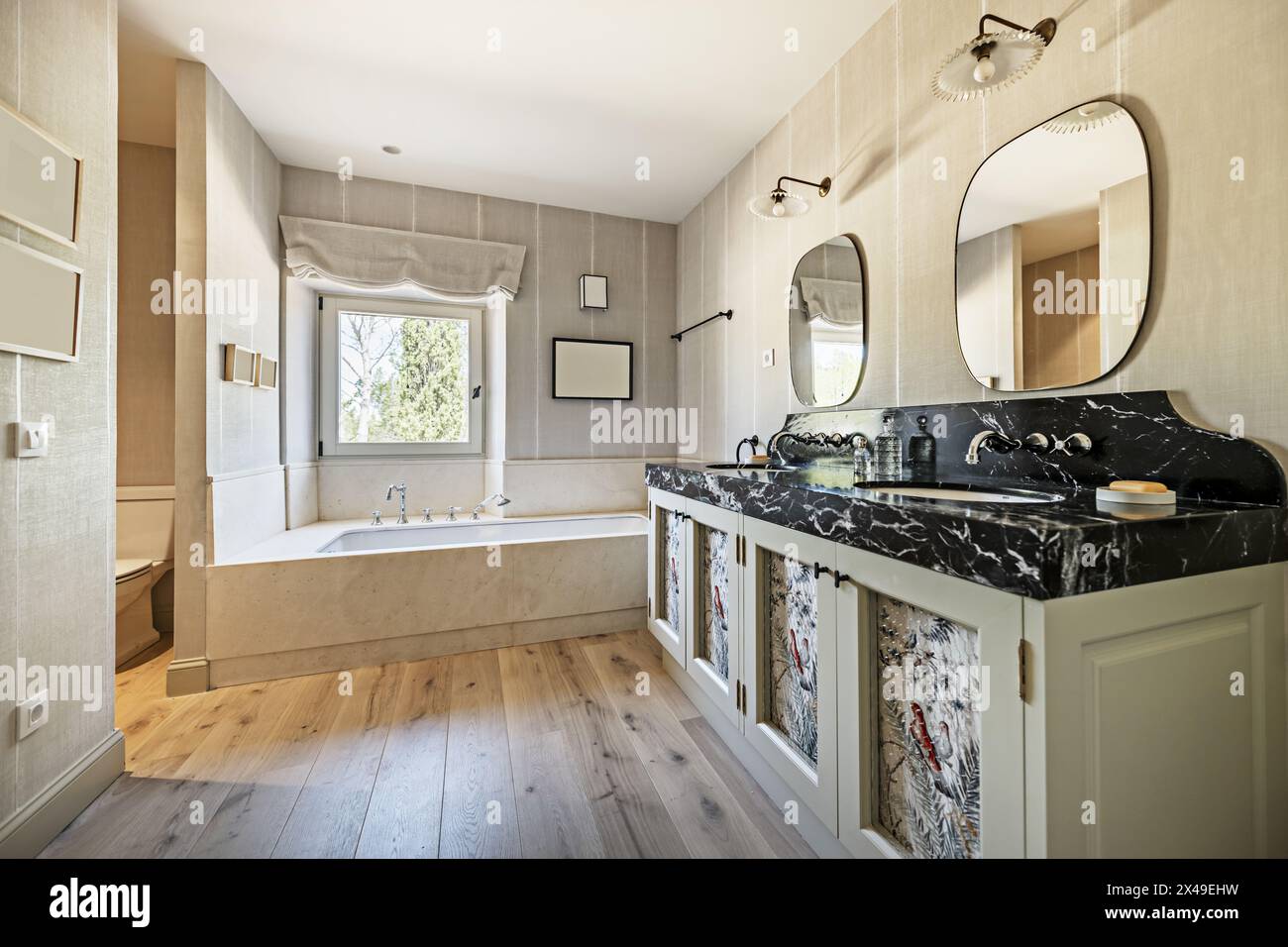 Un ampio bagno con mobili in legno con doppio lavabo, marmo nero, due specchi e una vasca con rubinetti in stile vintage e windo bianco in alluminio Foto Stock