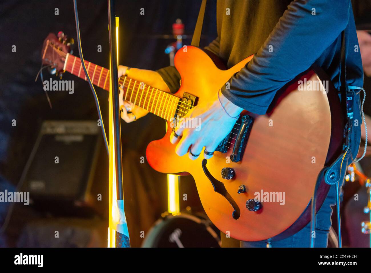 Un musicista sta suonando una chitarra elettrica semi-cava arancione sul palco con luci gialle luminose sullo sfondo. Il chitarrista sta sta indossando un blac Foto Stock