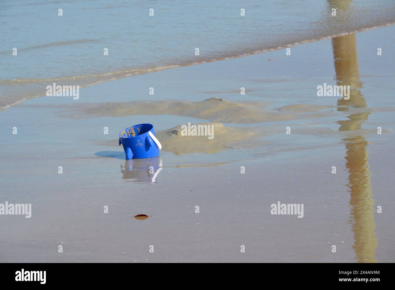 Un secchio blu abbandonato riposa, mezzo sommerso nella sabbia vicino al bordo dell'oceano, mentre le onde si innalzano, creando riflessi sulla sabbia delle spiagge. Foto Stock