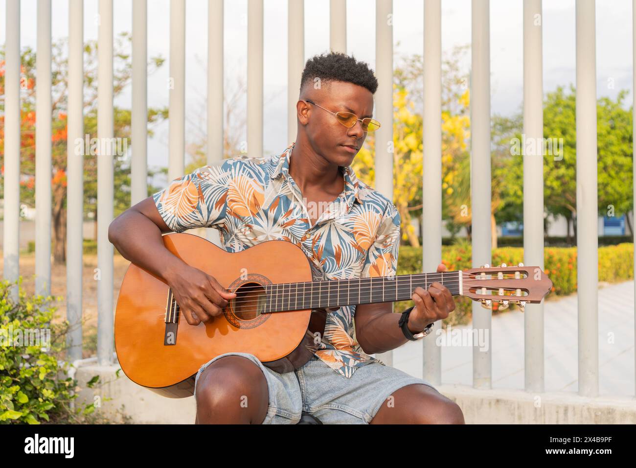 Un uomo africano con occhiali da sole e una camicia floreale suona la chitarra, circondato da un giardino vivace e da un'architettura moderna. Foto Stock