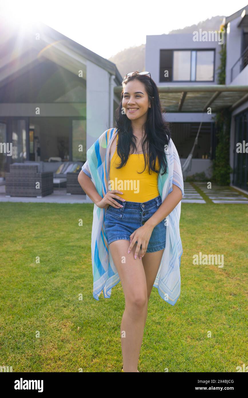 Giovane donna birazziale in piedi all'aperto in un cortile soleggiato. Indossando un top giallo e pantaloncini in denim, tenendo lo scialle leggero, godendosi la calda luce del sole, inalterato Foto Stock