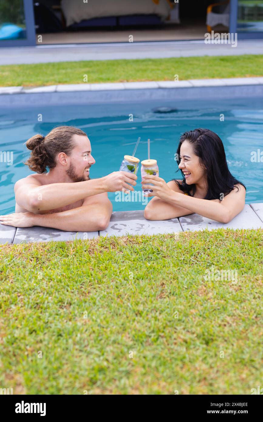 Coppie diverse che si godono un drink all'aperto vicino alla piscina, ridendo insieme. Lei, con i capelli scuri, e lui, con i lunghi capelli castani e la barba, si stanno divertendo Foto Stock