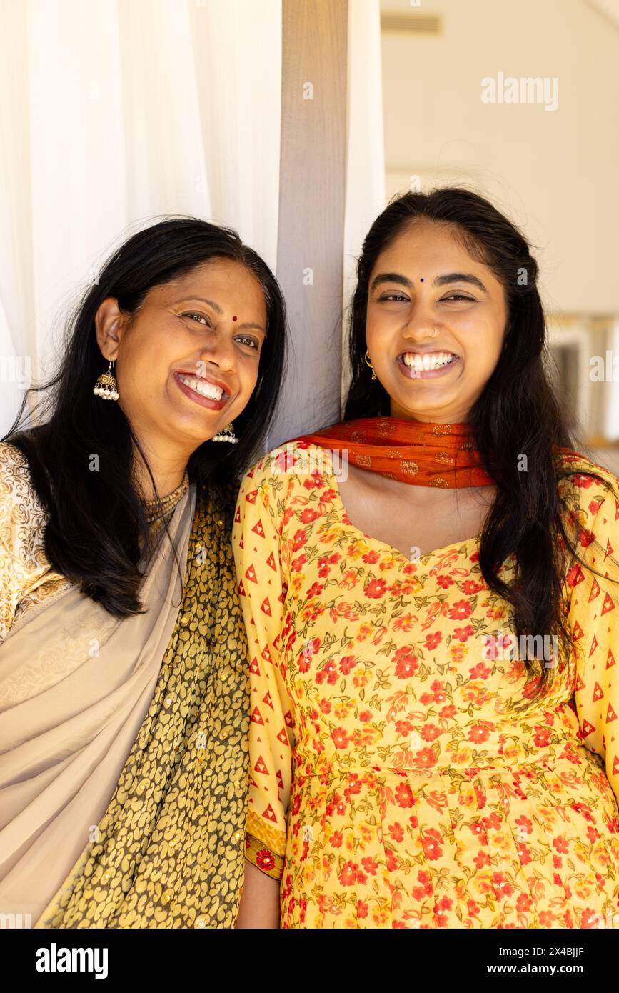 Madre indiana e figlia adolescente, entrambe sorridenti, in piedi insieme a casa. Madre, mostra striature di grigio nei suoi capelli scuri, tenendosi per mano Foto Stock