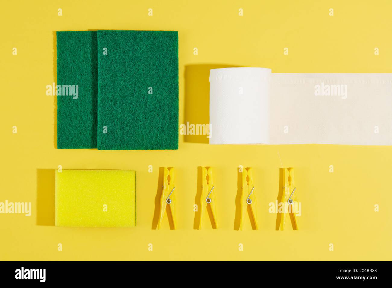 Materiali per la pulizia - spugne verdi, rotolo di carta bianca e clothespins decorati su sfondo giallo. Vista dall'alto, scena minima per la pulizia della pubblicità Foto Stock