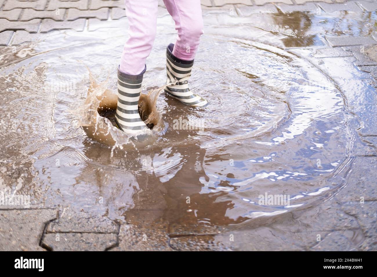 la maliziosa ragazza di 5 anni con stivali di gomma salta con gioia in una pozza, ravvicinare i piedi dei bambini in schizzi d'acqua, catturando le gioie pure e semplici del bambino Foto Stock