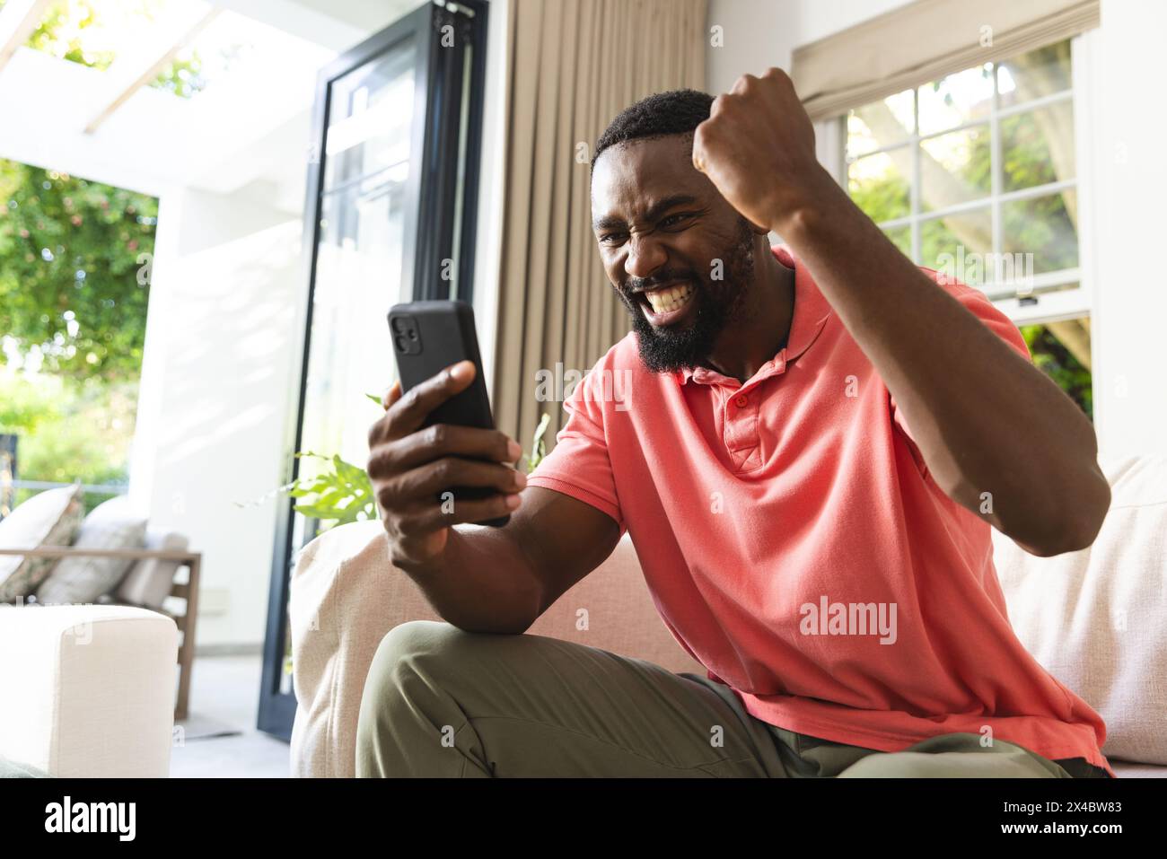 Uomo afroamericano che festeggia mentre guarda lo smartphone a casa. Indossa una camicia rosa, ha i capelli neri corti e una barba, inalterata. Foto Stock