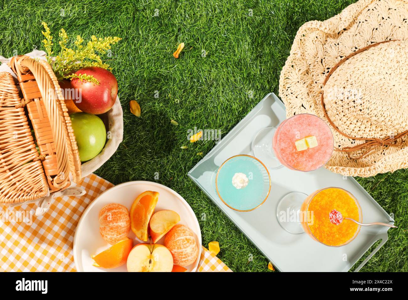 Idea estiva con set da picnic decorato su erba verde. Cestino e piatto pieno di frutta, tazze di frullati freschi e cappello a teglia decorato sul dorso dell'erba Foto Stock