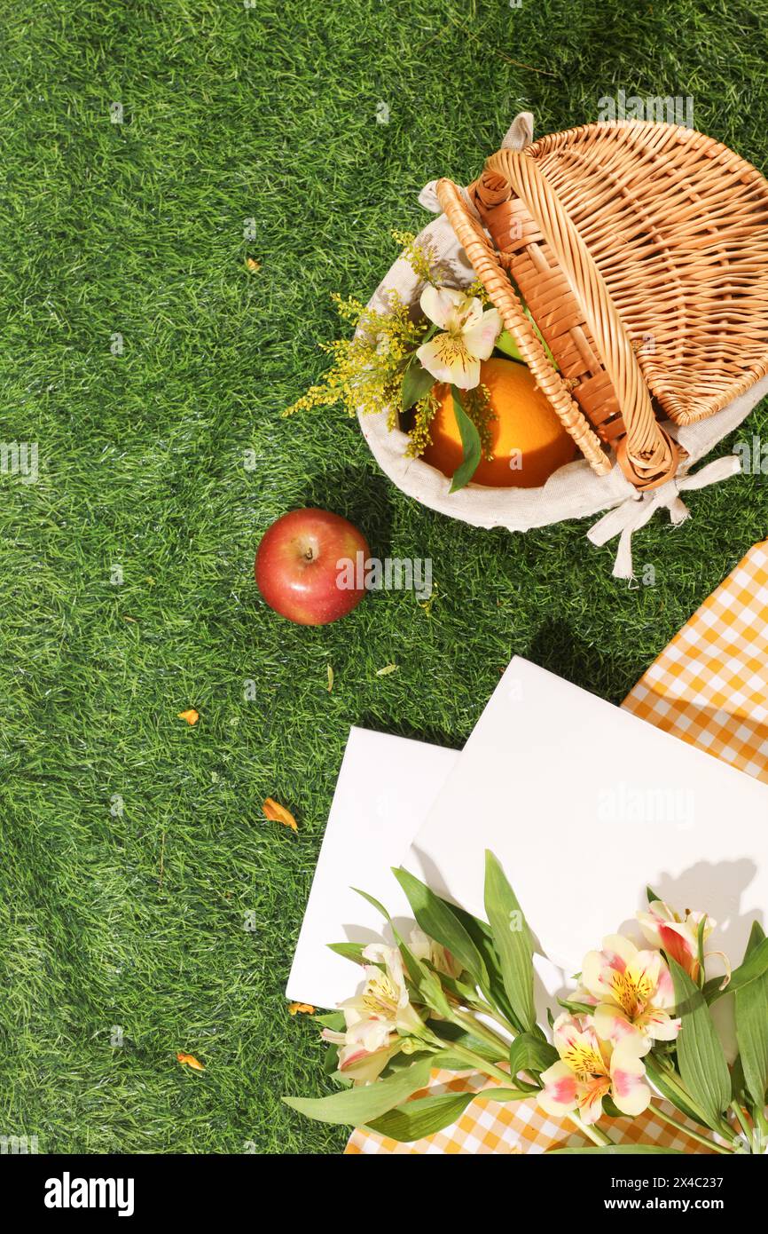 Un cestino di bambù con frutta e fiori decorato con quaderni bianchi e tessuto a scacchi su sfondo verde. Vista dall'alto, spazio di copia per Foto Stock