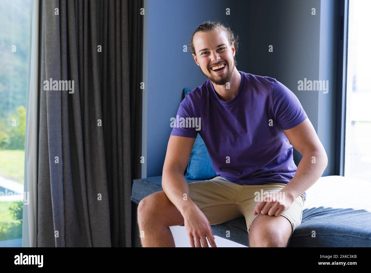 Giovane caucasico seduto, sorridente, con abiti casual a casa, spazio fotocopie. Ha la barba, i capelli lunghi legati, ed è in una stanza ben illuminata, unal Foto Stock