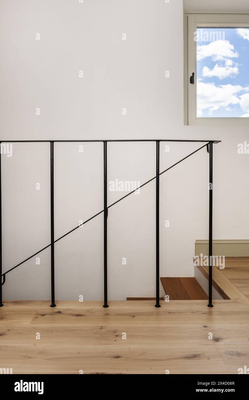 Scala con gradini in legno di quercia di una casa indipendente con ringhiera in metallo di stile minimalista nero, semplici pareti bianche e finestre quadrate Foto Stock