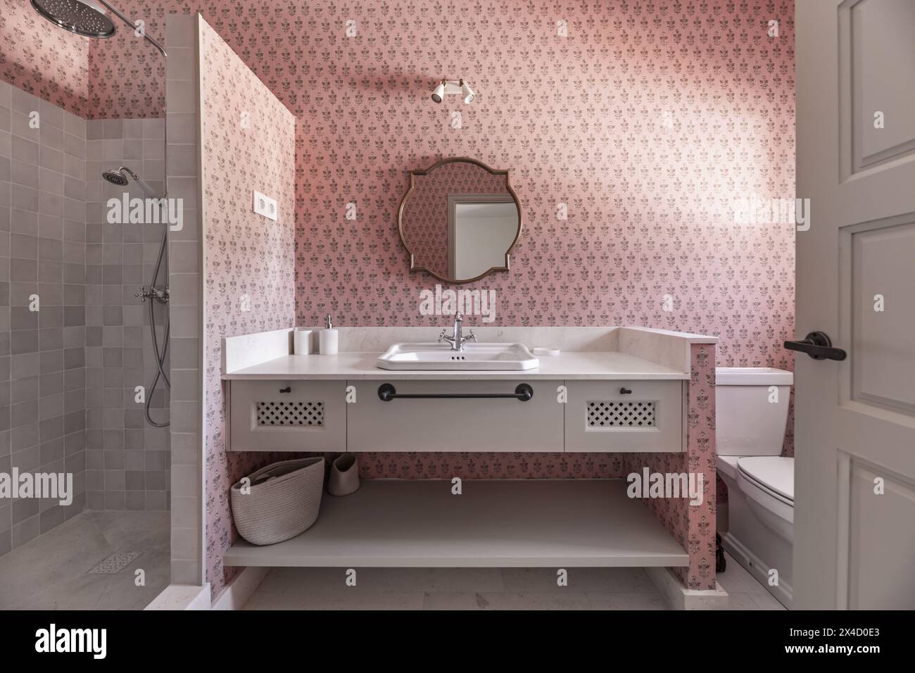 Un bellissimo bagno dal design moderno con lavandino in marmo color crema, specchio incorniciato in legno dorato, mobili e pareti coordinati con carta da parati rosa decorativa Foto Stock