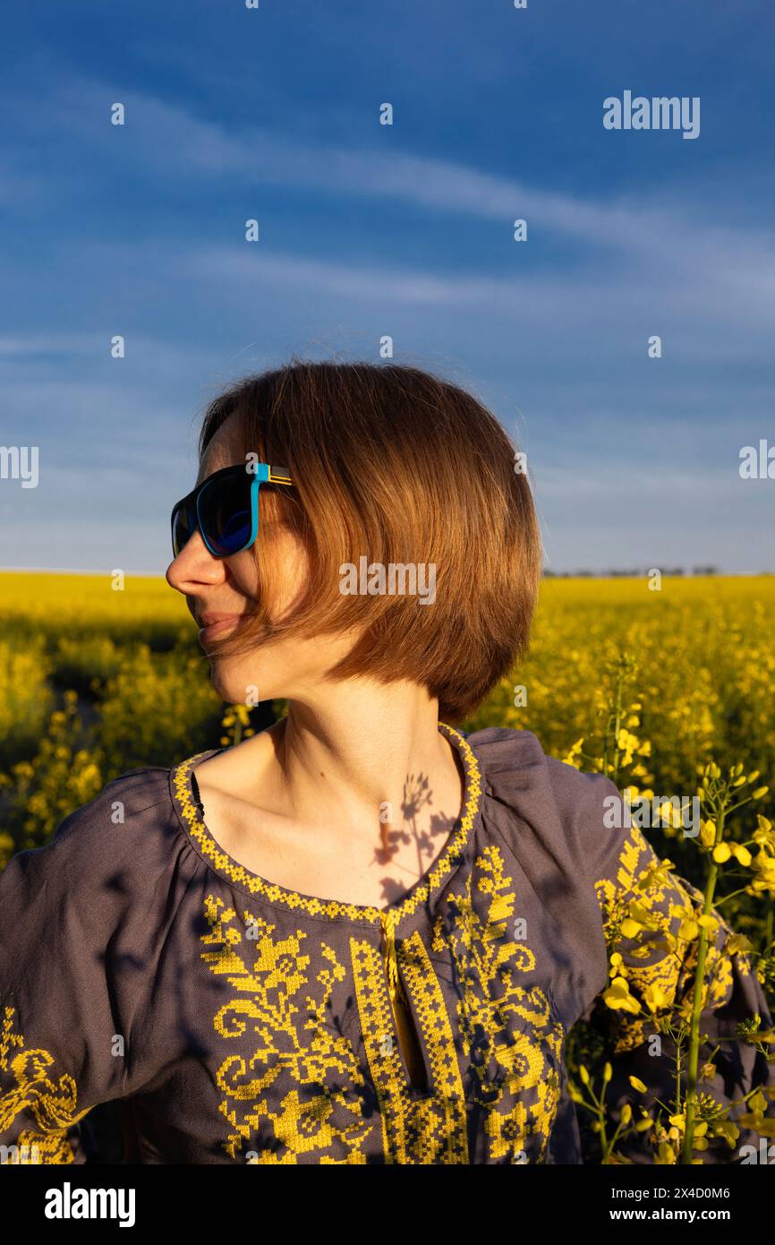 Donna felice di 35-40 anni con occhiali da sole blu e camicetta ricamata in Ucraina in un campo di colza. detox digitale, weekend estivi, goditi il momento Foto Stock