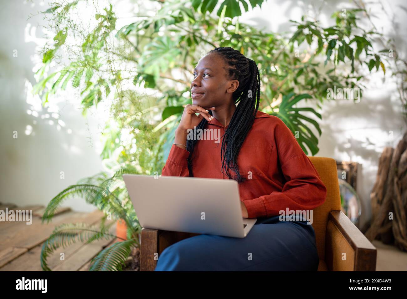 Una donna nera premurosa e rilassata si prende una pausa dal lavoro con un laptop, guarda da parte la finestra e la verde casa. Foto Stock