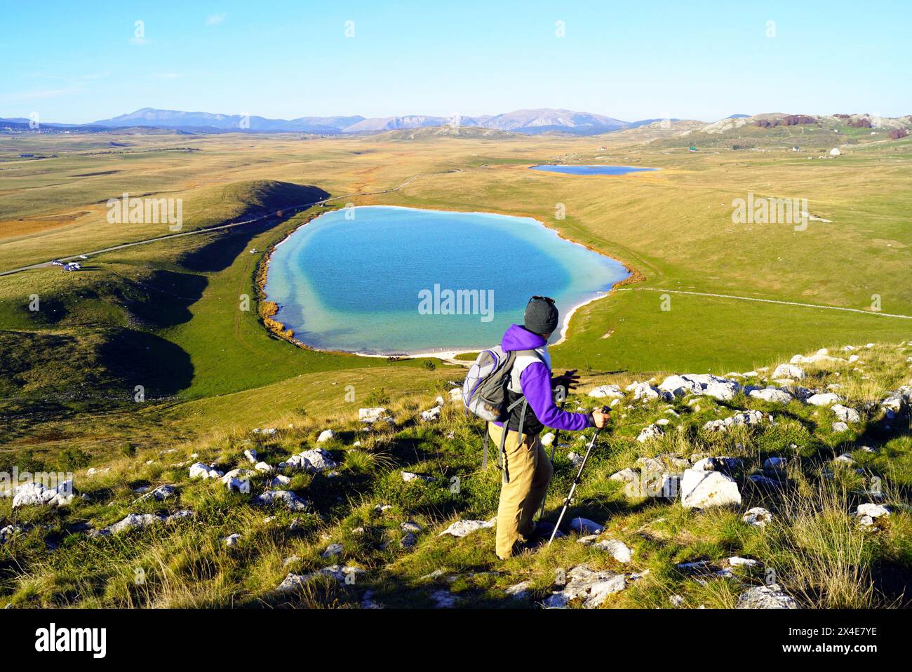 Un escursionista con bastoni da trekking si trova di fronte a un bellissimo lago blu nel mezzo di una pianura erbosa. Vacanze attive in Montenegro Foto Stock