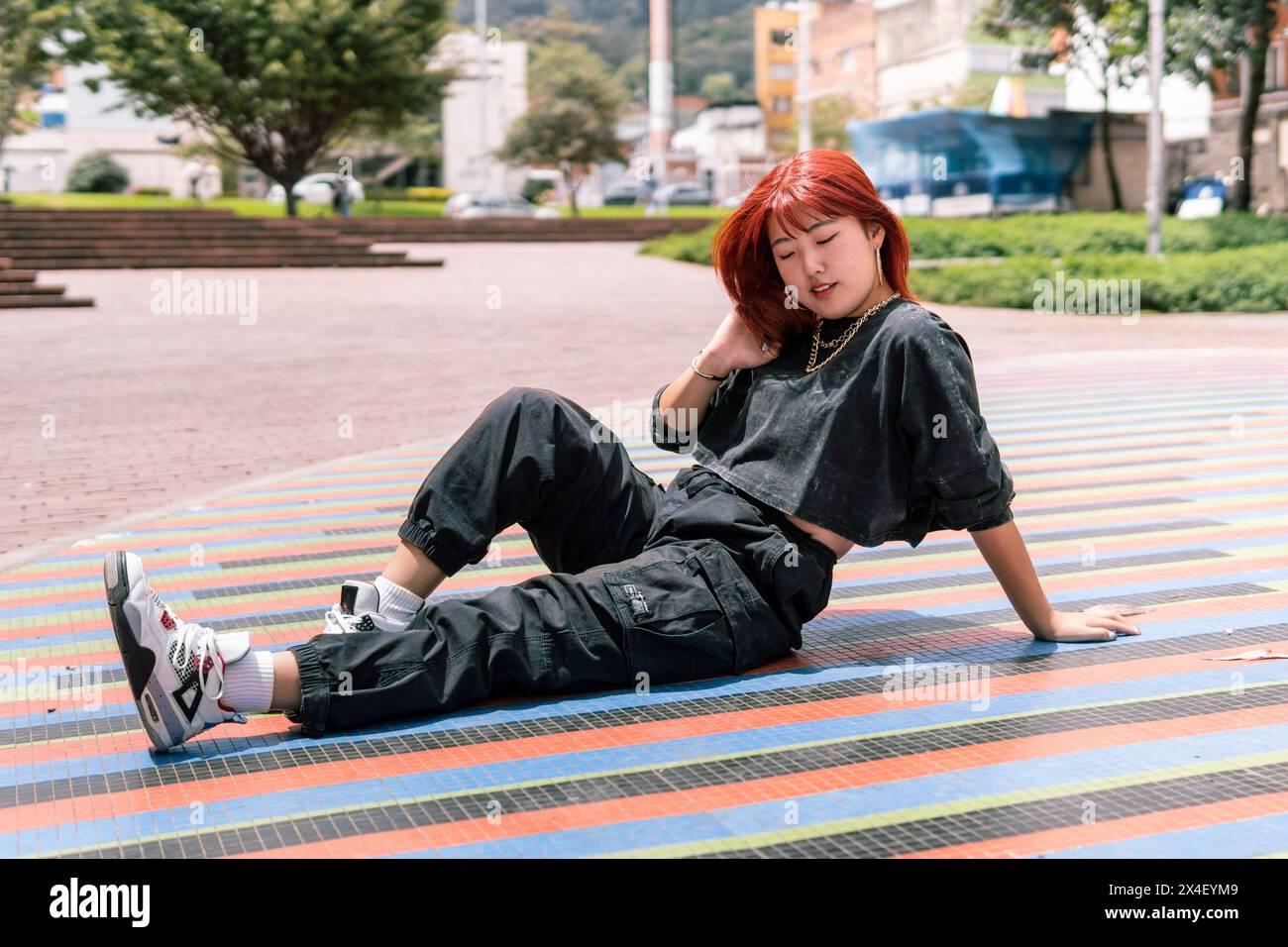 Elegante donna coreana con capelli rossi che si rilassa su un vivace mosaico in un parco urbano Foto Stock