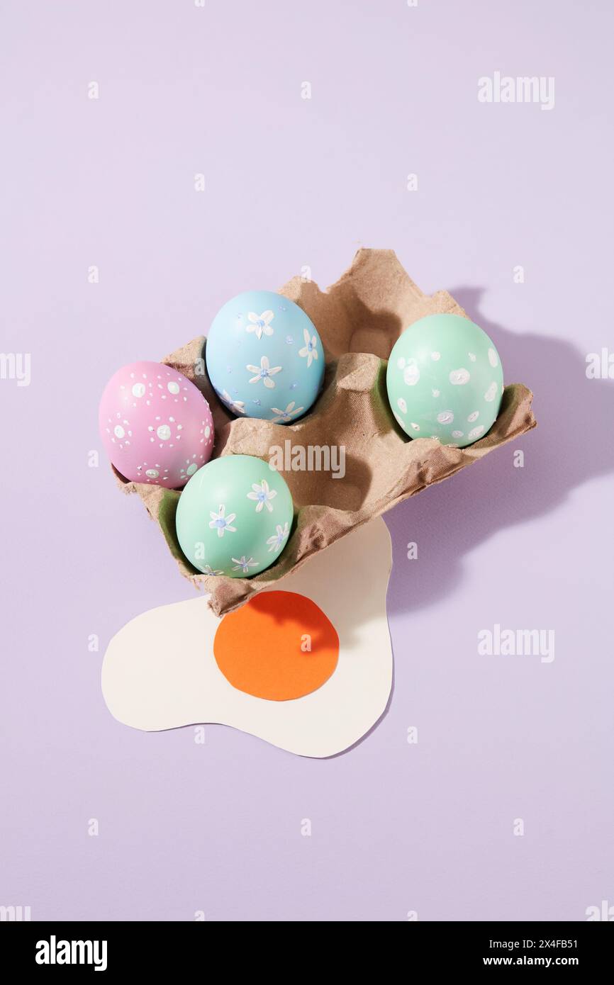 Vista dall'alto di un piccolo blister con uova di Pasqua pastello e uova fritte decorate su sfondo viola. Scena minimal con un regalo significativo per il giorno di Pasqua Foto Stock