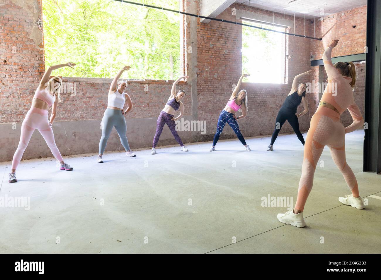 Questa immagine cattura un momento di esercizio collettivo in cui un gruppo di donne partecipa a una routine di stretching. I loro movimenti sono in armonia, creando un senso di unità e sincronizzazione. La spaziosa palestra in stile industriale con i suoi mattoni a vista e le grandi finestre offre uno spazio luminoso e arioso per le loro attività fitness. L'abbigliamento femminile varia, riflettendo stili personali, ma il loro sforzo coordinato indica un obiettivo di fitness condiviso. Movimento armonizzato: Sessione di stretching di gruppo. Foto di alta qualità Foto Stock