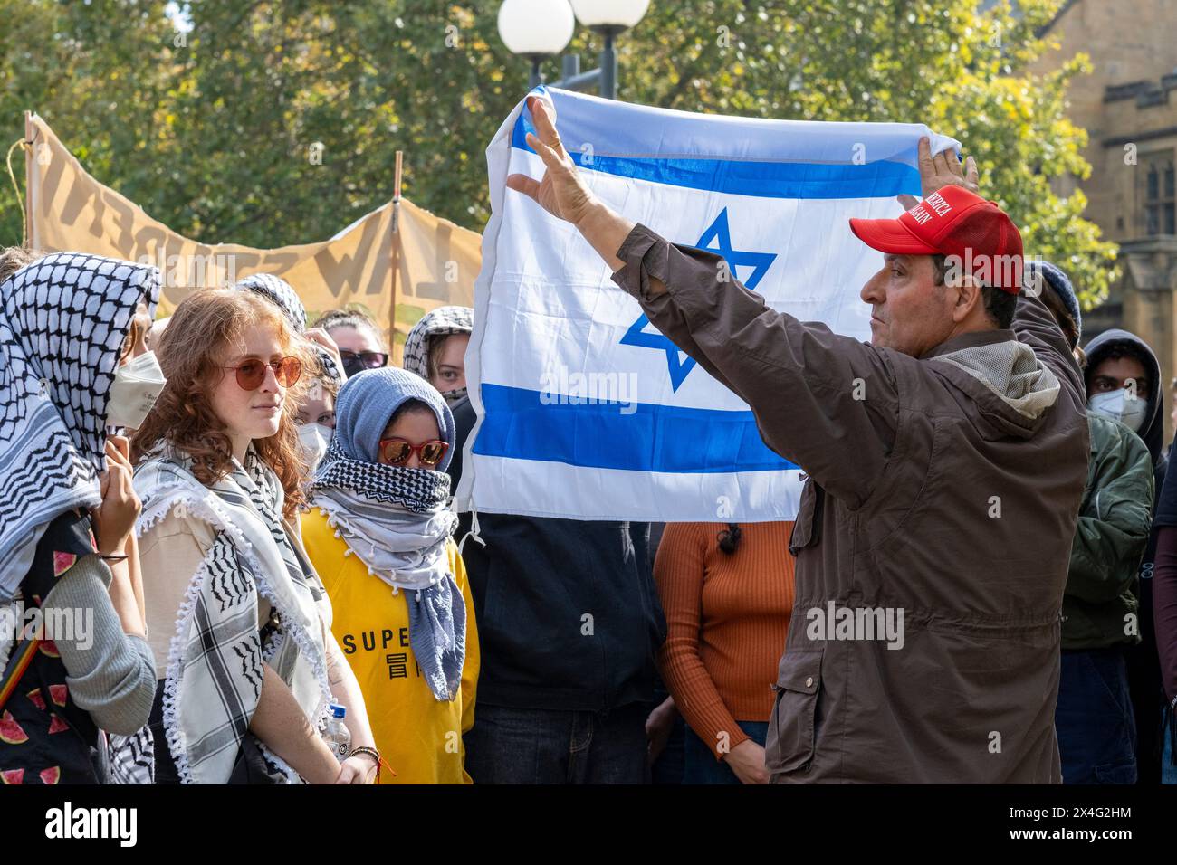 Un contro-manifestante inonda una bandiera israeliana contro i manifestanti durante una manifestazione filo-palestinese nel campus dell'Università di Melbourne. Il pro-israeliano sta indossando un berretto Make America Great Again. Melbourne, Victoria, Australia. Foto Stock