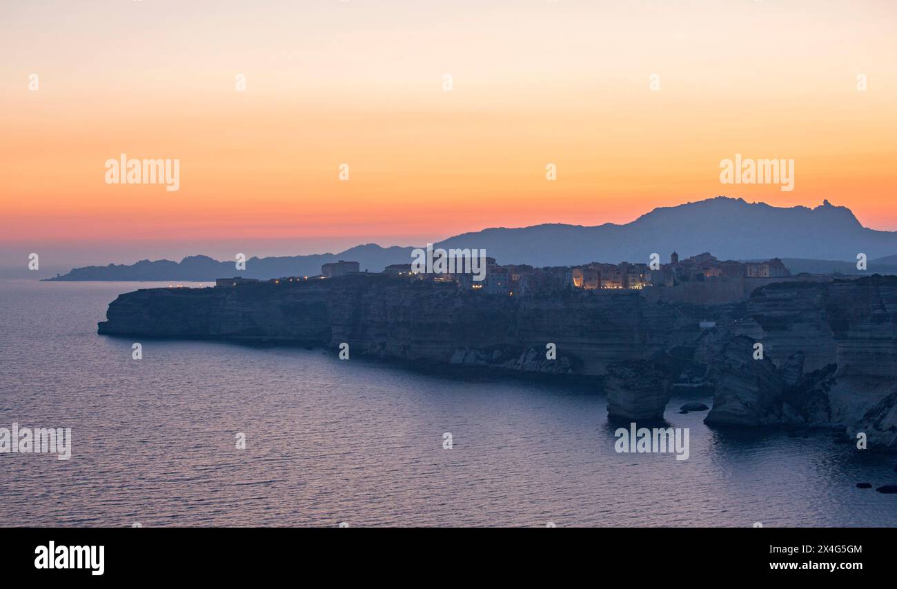 Bonifacio, Corse-du-Sud, Corsica, Francia. Ammira dalla cima della scogliera la cittadella illuminata, il crepuscolo, le montagne lontane che si stagliano contro un cielo dorato. Foto Stock