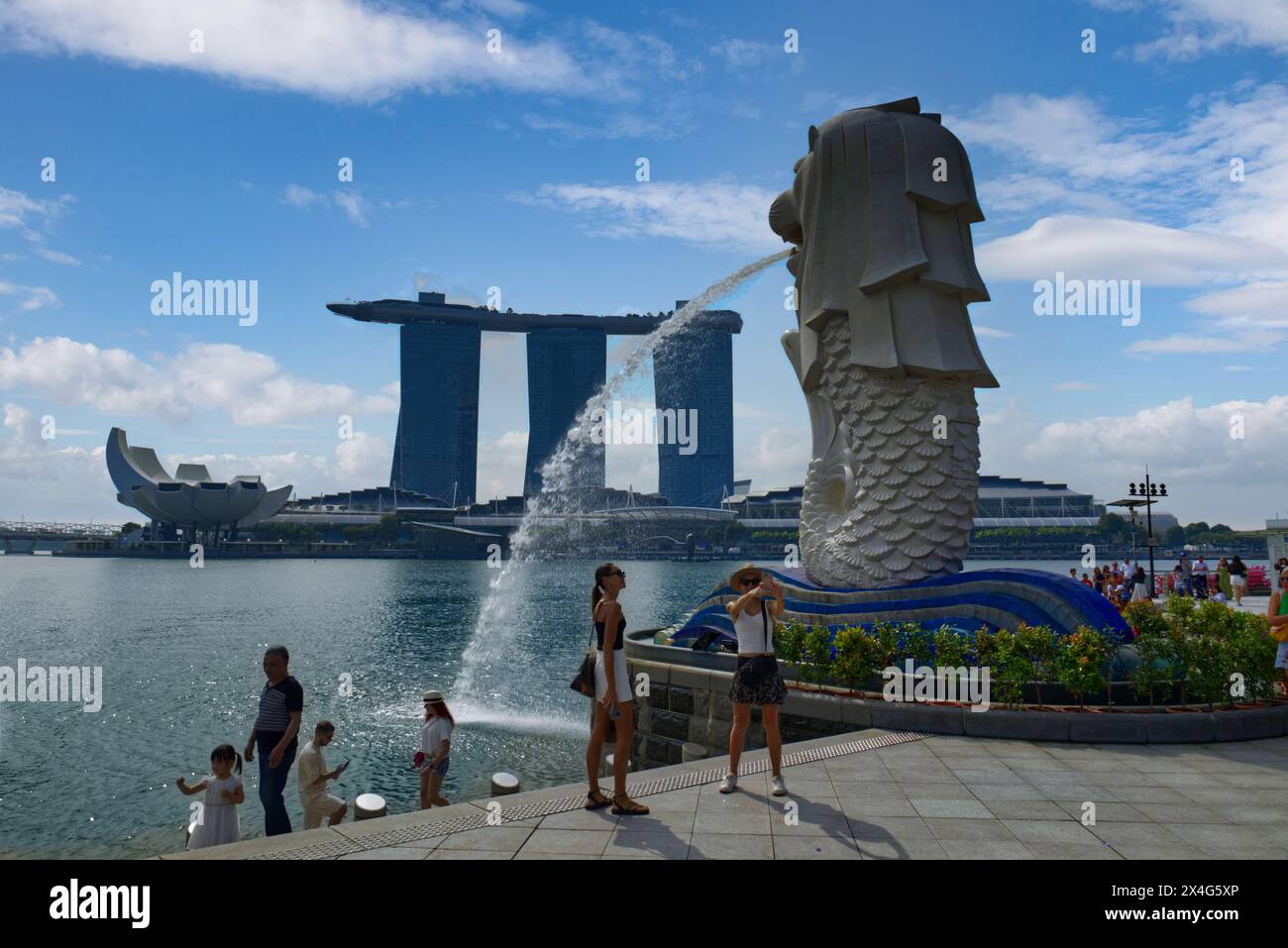 Persone che si godono il Merlion Park, il punto di riferimento di Singapore e l'attrazione turistica con la fontana della testa del leone sullo sfondo del resort Marina Bay Sands Foto Stock
