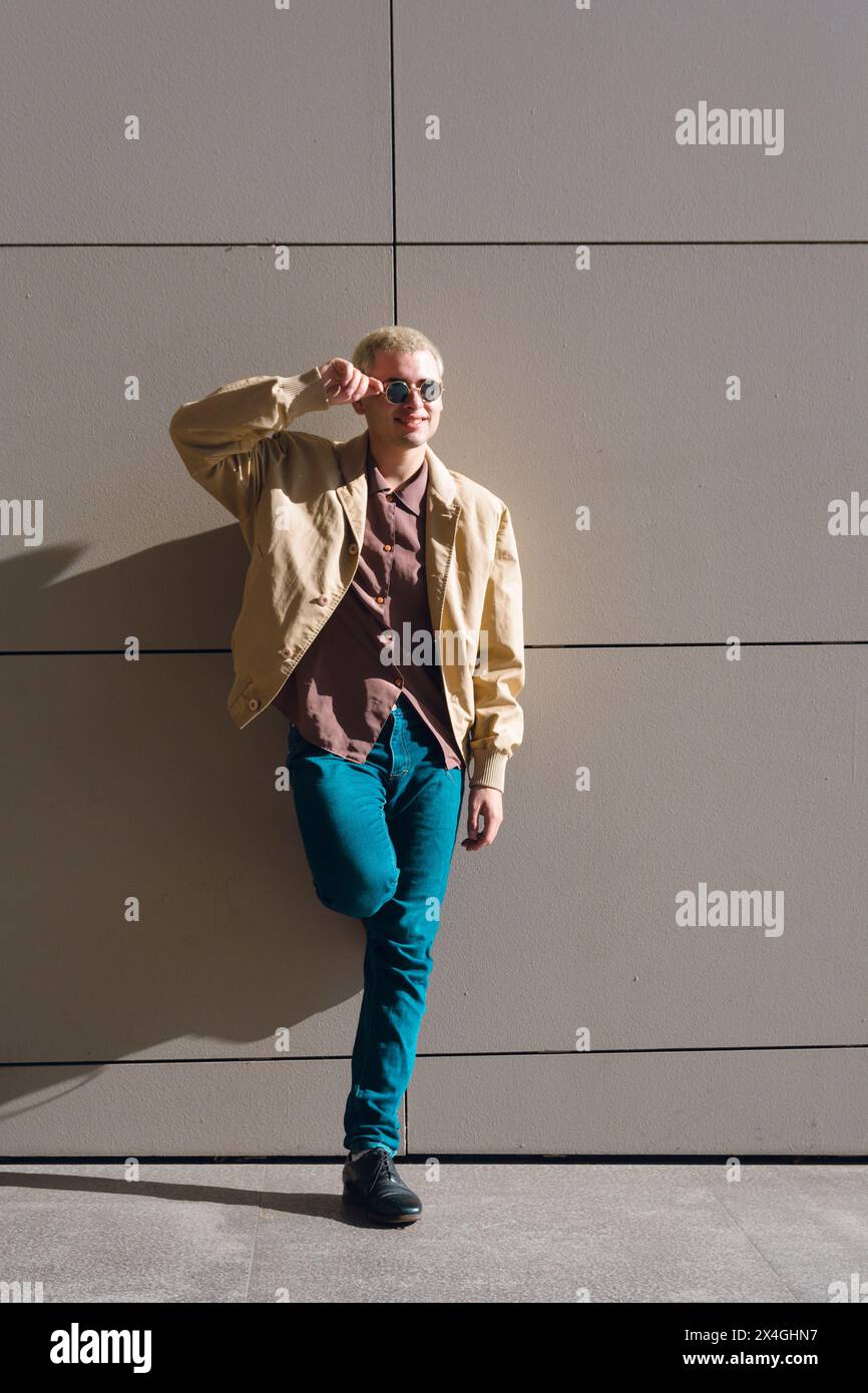 giovane gay con occhiali in piedi contro il muro al tramonto, con giacca marrone e pantaloni jean Foto Stock