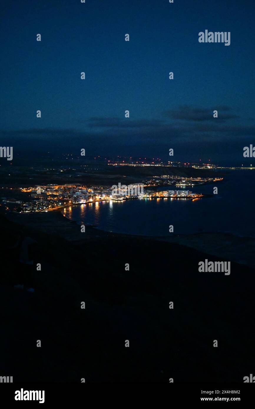 le luci notturne della città riflettono le acque costiere, creando uno scintillante arazzo di radiosità urbana contro il cielo notturno blu profondo, un panorama mozzafiato Foto Stock