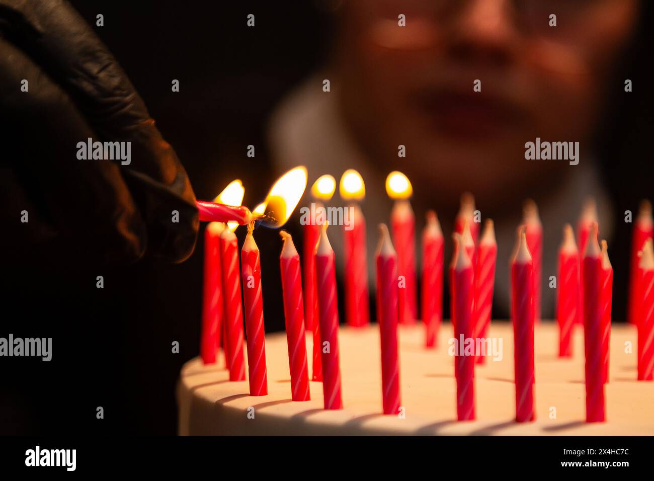 Goiania, Goias, Brasile – 27 aprile 2024: Dettaglio di una mano con un guanto nero, che accende diverse candele su una torta di compleanno con un volto sfocato. Foto Stock