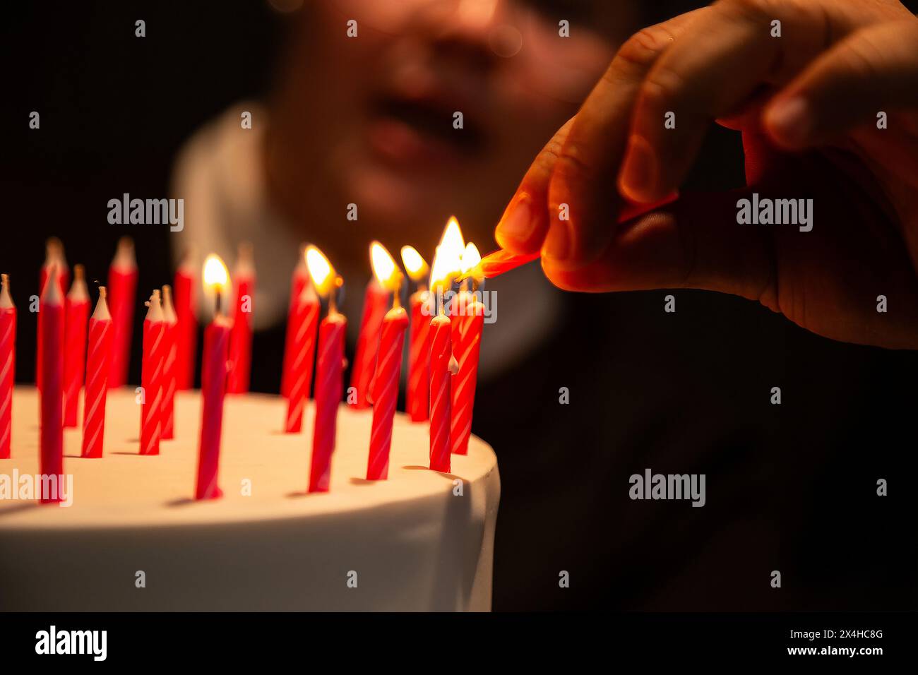 Goiania, Goias, Brasile – 27 aprile 2024: Dettaglio di una mano che accende diverse candele su una torta di compleanno con un volto sfocato sullo sfondo. Foto Stock