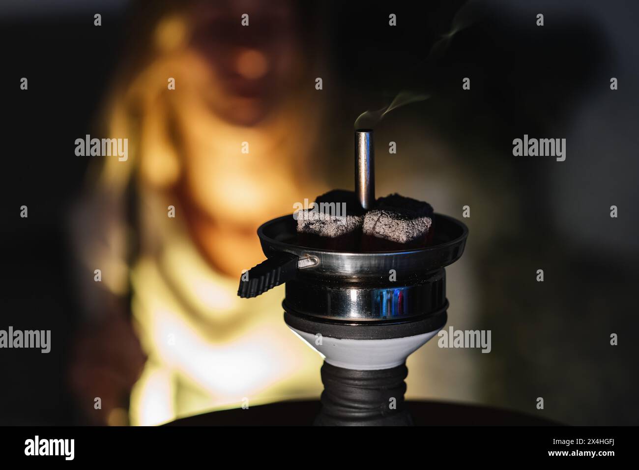 Primo piano di carboni scintillanti su un narghilè con fumo, donna sfocata sullo sfondo, sotto la luce calda del sole in una tradizionale lounge di narghilè araba. Foto Stock