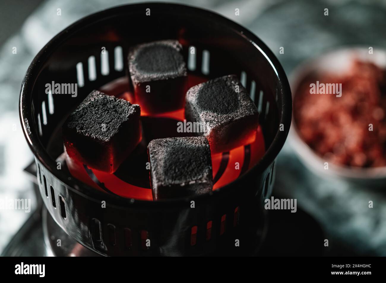 Carboni accesi riscaldano in un accendino elettrico a carbone per shisha, con una ciotola di tabacco sfocata sullo sfondo in una tradizionale lounge di narghilè araba. Foto Stock