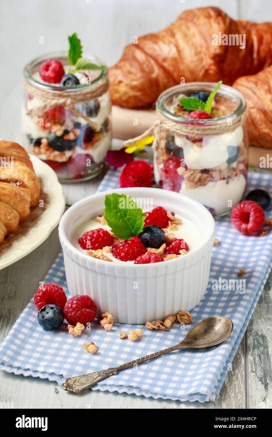Tavolo della colazione: Ciotola di yogurt con muesli e frutta fresca: Lamponi e mirtilli, vasetti di vetro con frutta e panna montata. Foto Stock