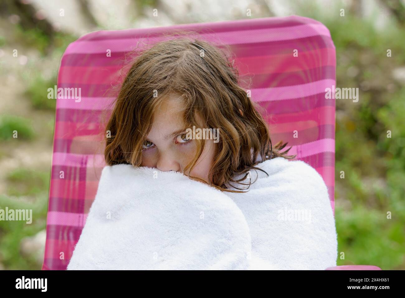 Bella bambina di otto anni seduta su una sedia avvolta in un asciugamano bianco dopo essere uscita dall'acqua, sembra fredda Foto Stock