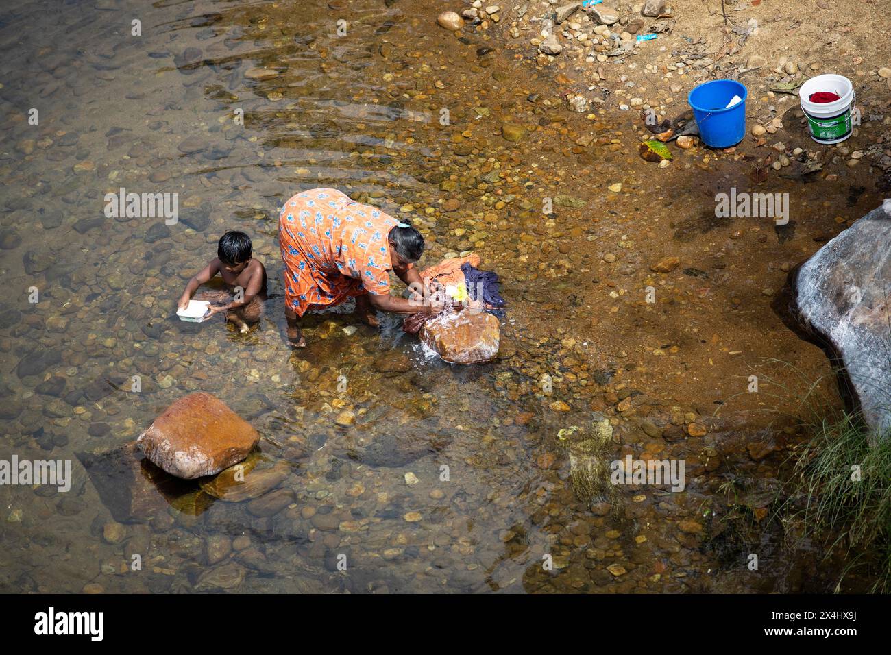 Donna indiana che lava vestiti nel fiume Periyar, suo figlio che gioca accanto a lei, Mundakayam, Kerala, India Foto Stock